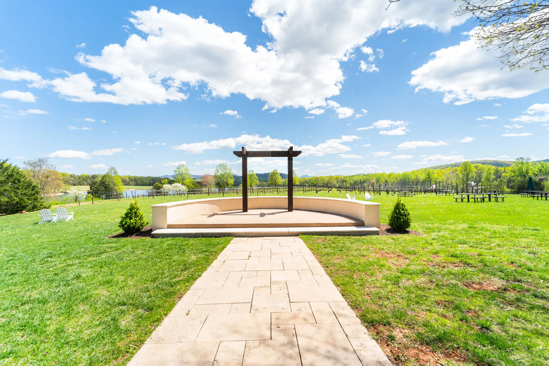 View of a grassy area with a stone path leading to a wooden arch overlooking a lake under a bright blue sky.