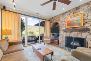 Living room with stone fireplace, large windows showcasing mountains, and wooden furniture.