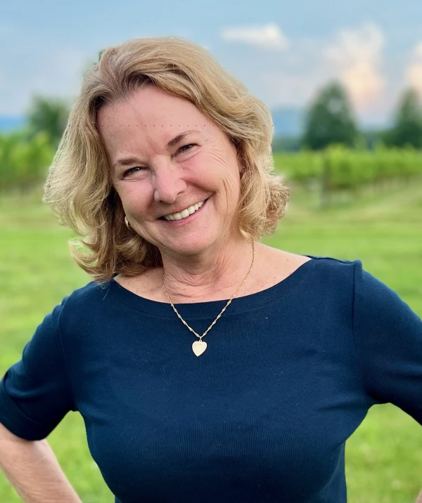 A photo of a smiling woman with blonde hair wearing a dark blue shirt in front of a vineyard background.