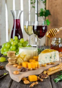 A wooden cheese board featuring a variety of cheeses, grapes, walnuts, and crackers, with honey being drizzled from a wooden dipper over a wedge of blue cheese, set against a background of wine glasses and a decanter.