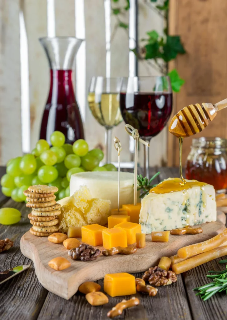 A wooden cheese board featuring a variety of cheeses, grapes, walnuts, and crackers, with honey being drizzled from a wooden dipper over a wedge of blue cheese, set against a background of wine glasses and a decanter.