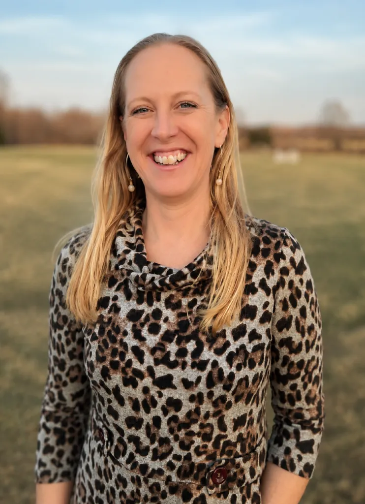Smiling woman with long blonde hair wearing a leopard print top standing outdoors in a grassy field.