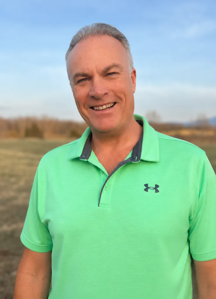 Smiling man wearing a bright green polo shirt standing outdoors in an open field at sunset.