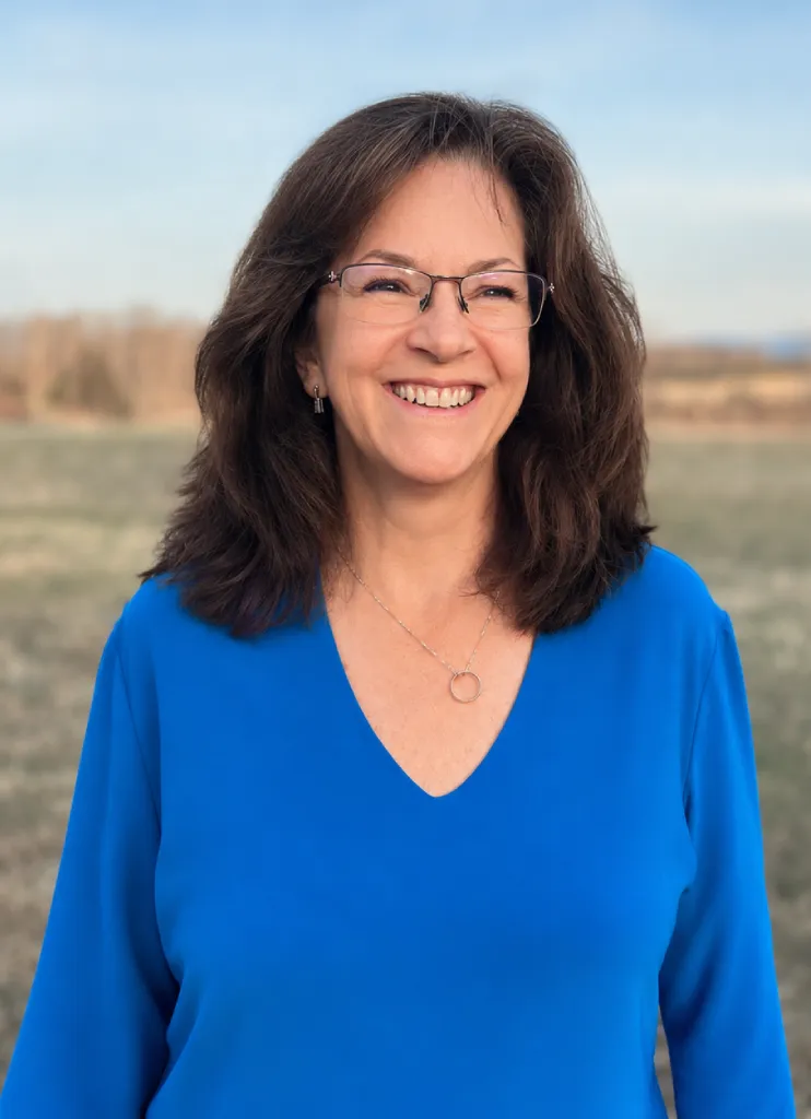 Smiling woman with glasses and dark hair wearing a bright blue blouse standing outdoors in a field.
