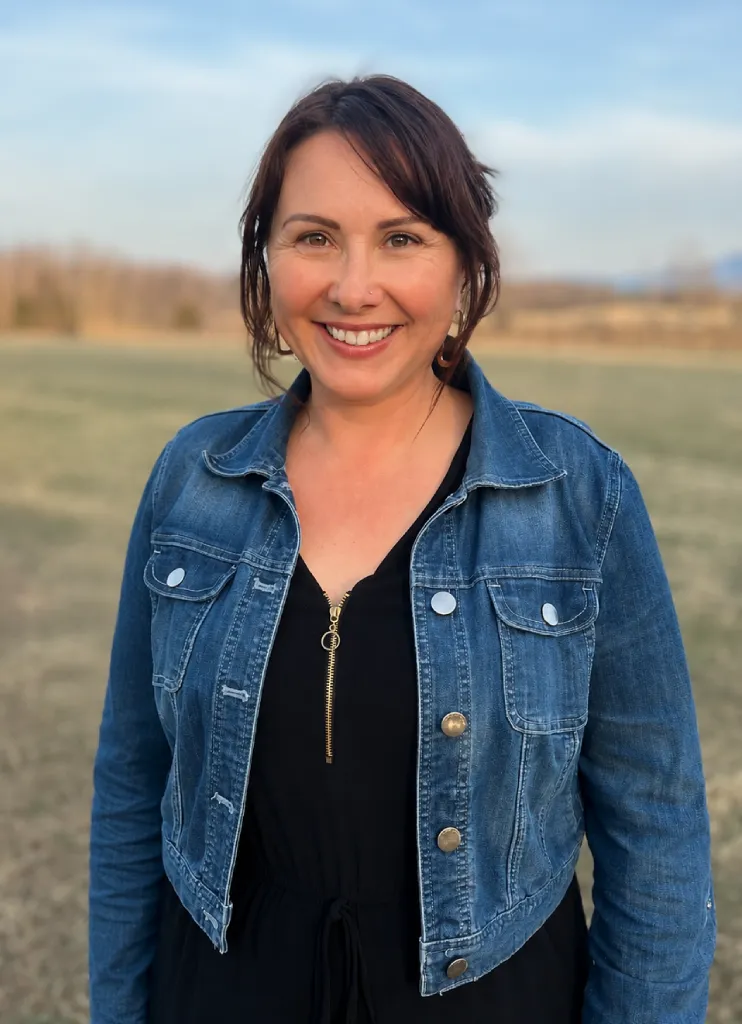 Smiling woman with dark hair wearing a denim jacket over a black dress standing outdoors in a grassy field.