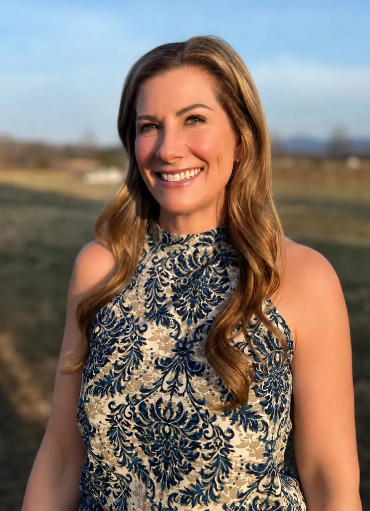 Smiling woman with long wavy hair wearing a sleeveless patterned dress standing outdoors during golden hour.