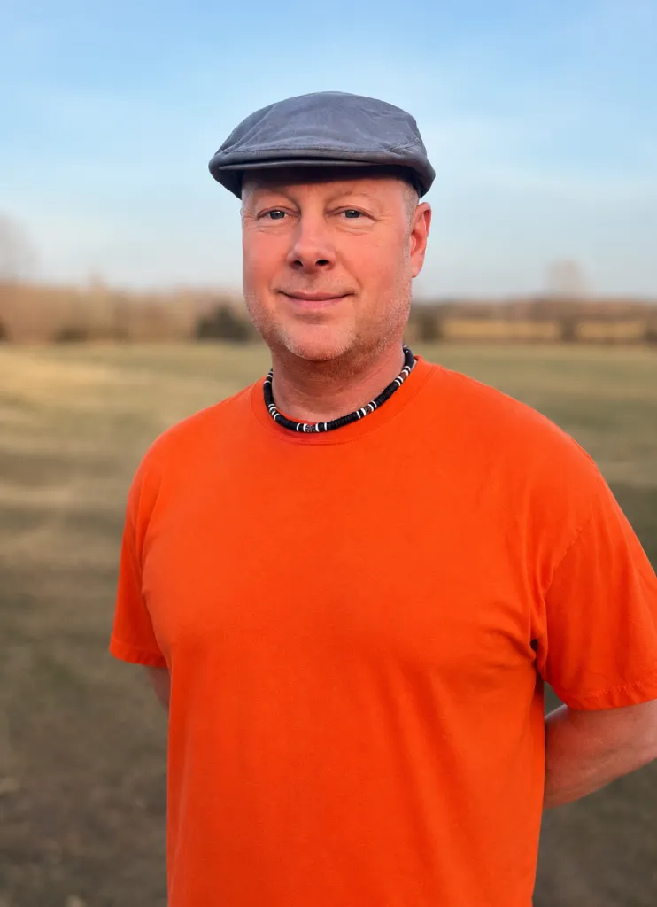 Man wearing an orange shirt and gray flat cap standing outdoors in an open field during sunset.