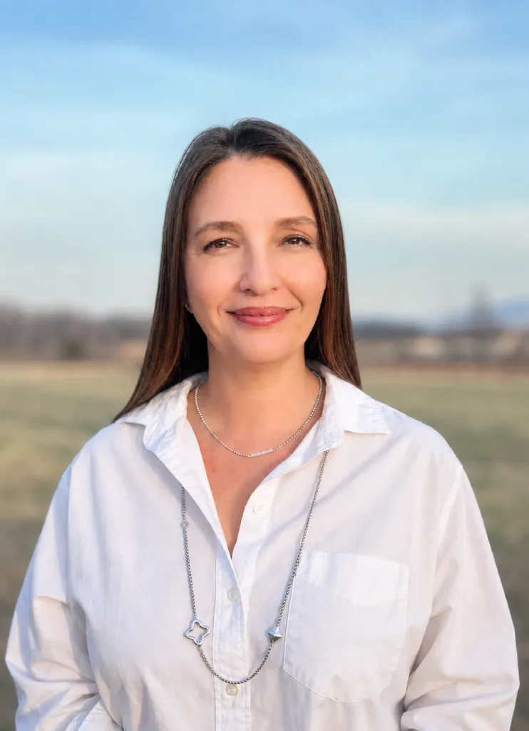 Smiling woman with long brown hair wearing a white blouse standing outdoors in a field.