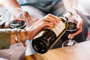 A close-up of hands pouring red wine from a bottle into a glass, with another hand holding a wine glass.