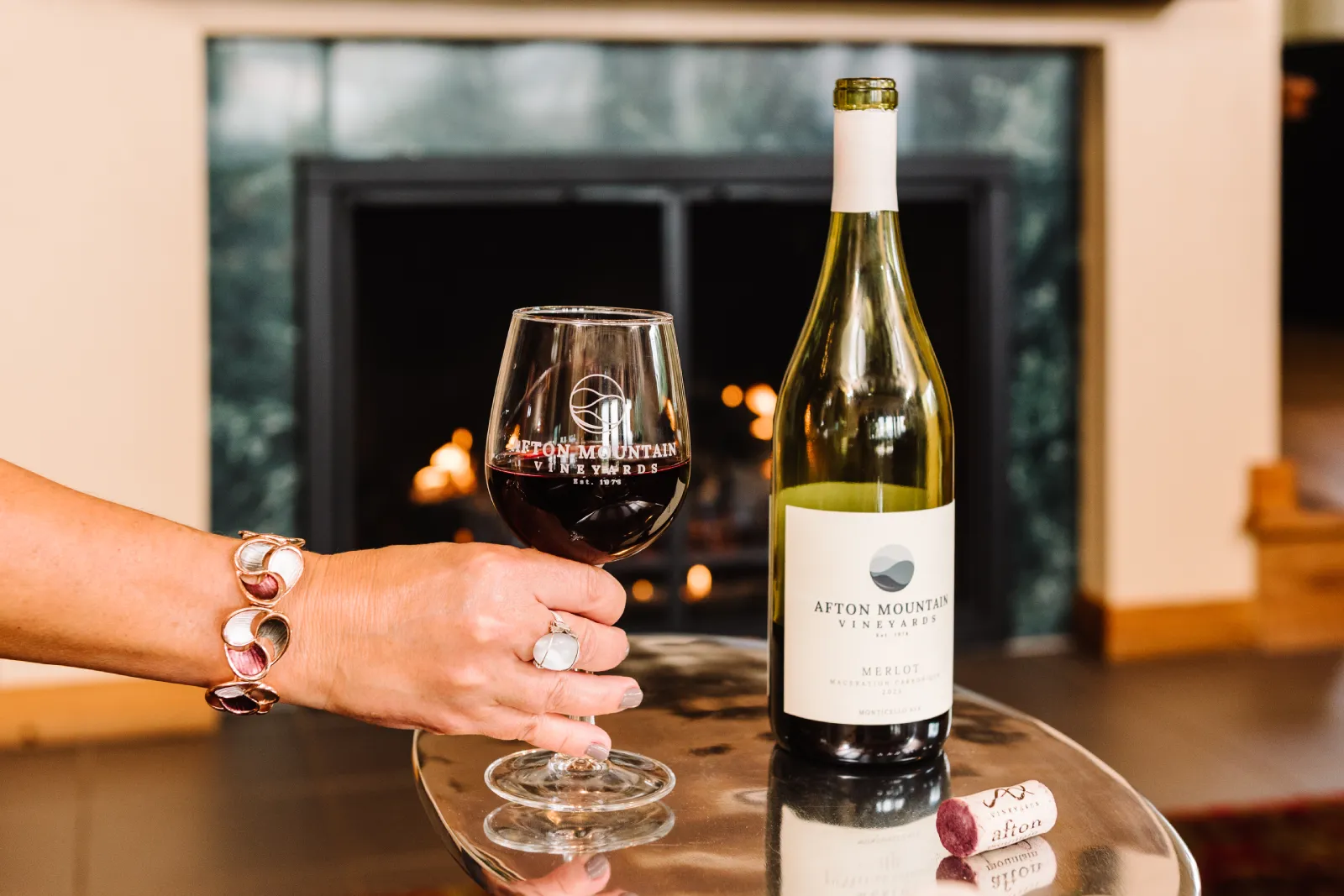 A glass of red wine on a tray with a bottle of Afton Mountain Vineyards Chardonnay and a cork. A cozy fireplace is in the background.